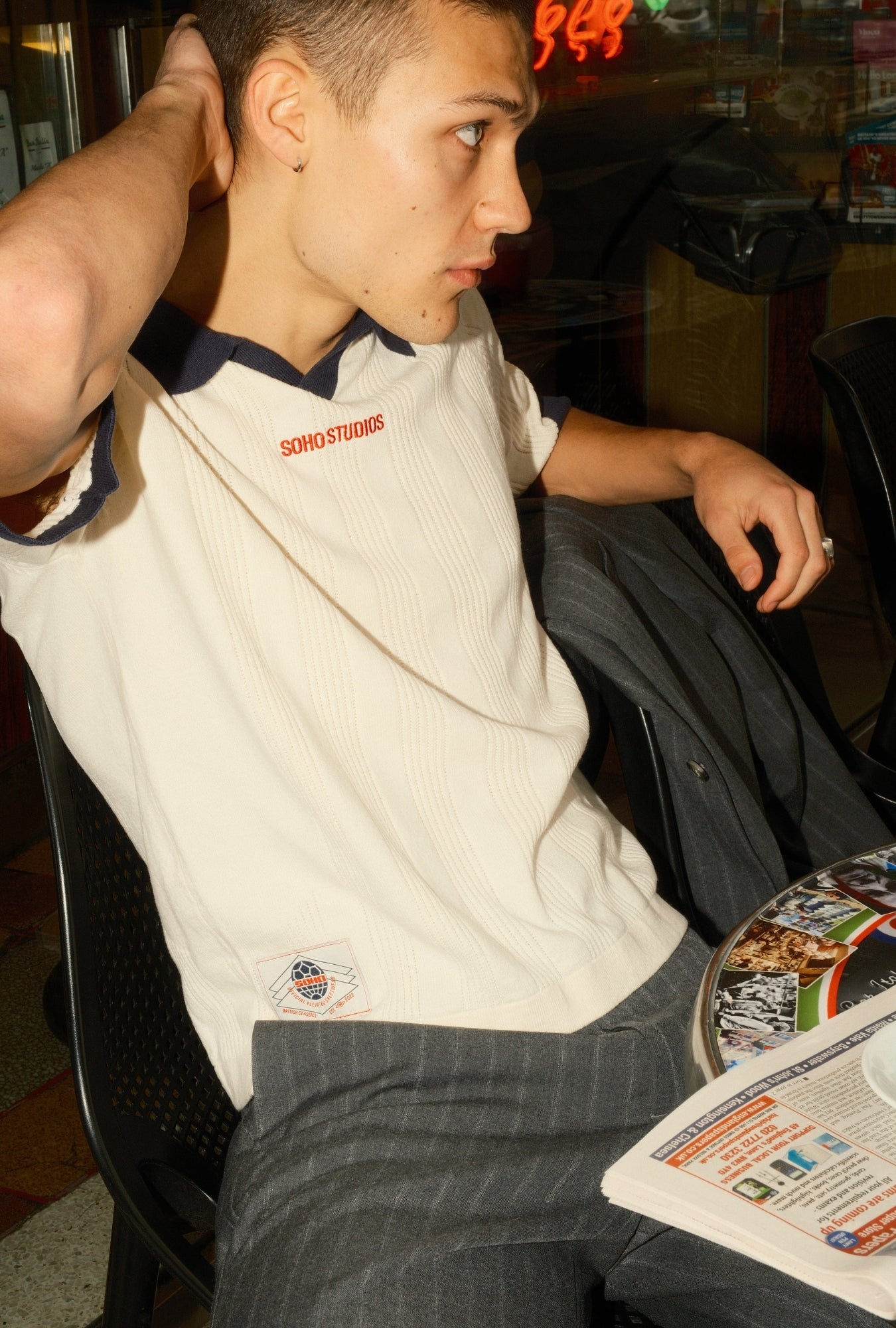 Man sitting at a table with a newspaper, wearing a white polo shirt with red text.