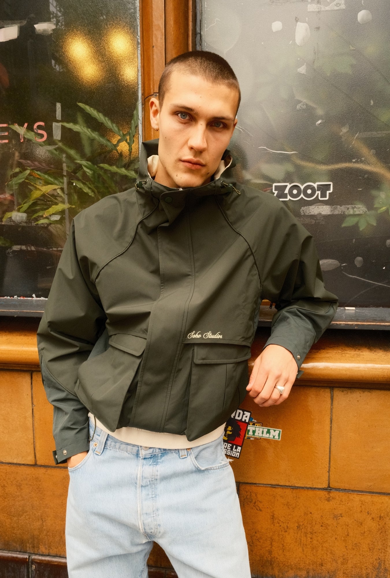 Man wearing a green jacket and light blue jeans standing indoors.