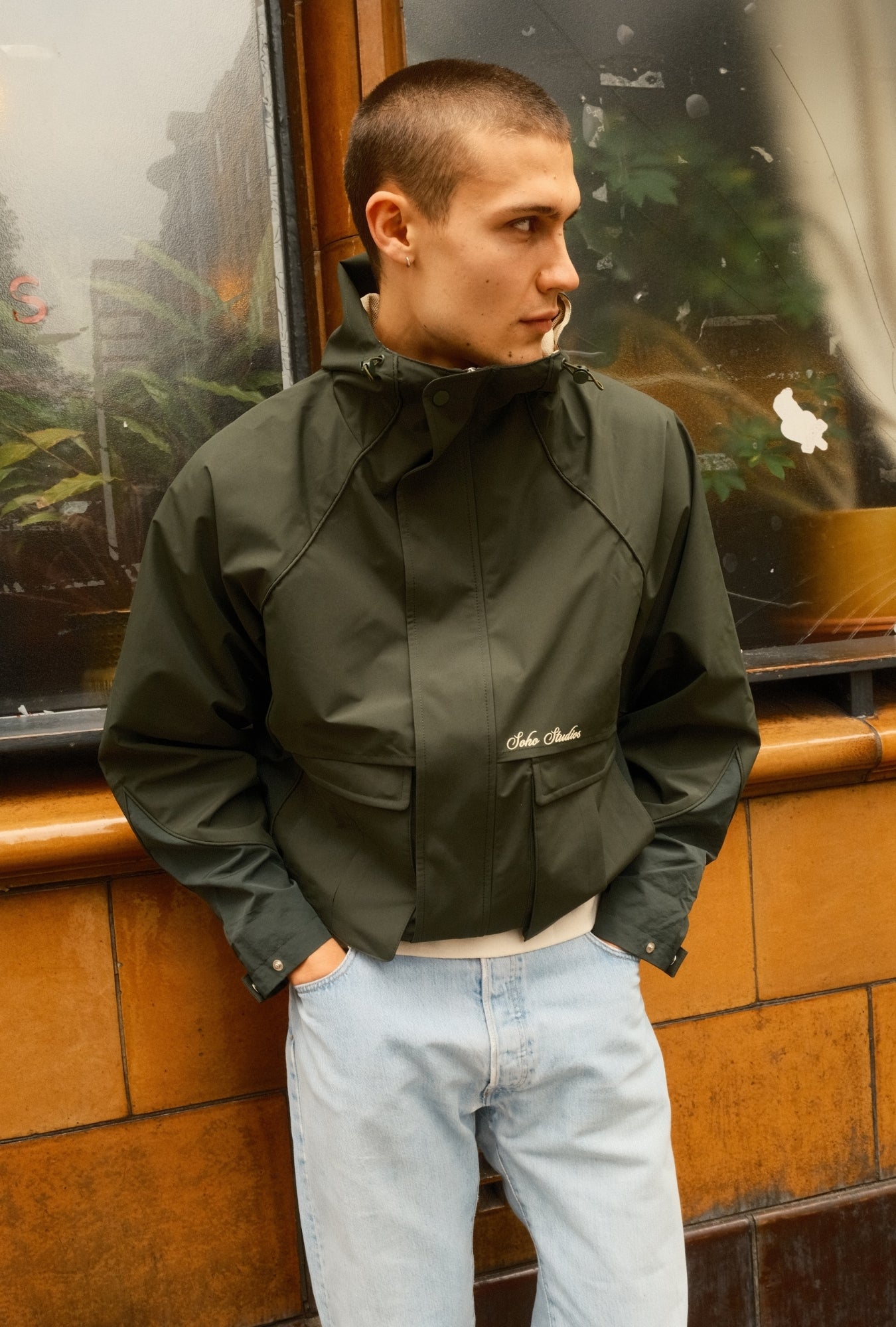 Person wearing a dark green jacket with a brand logo, standing against a reflective glass wall.