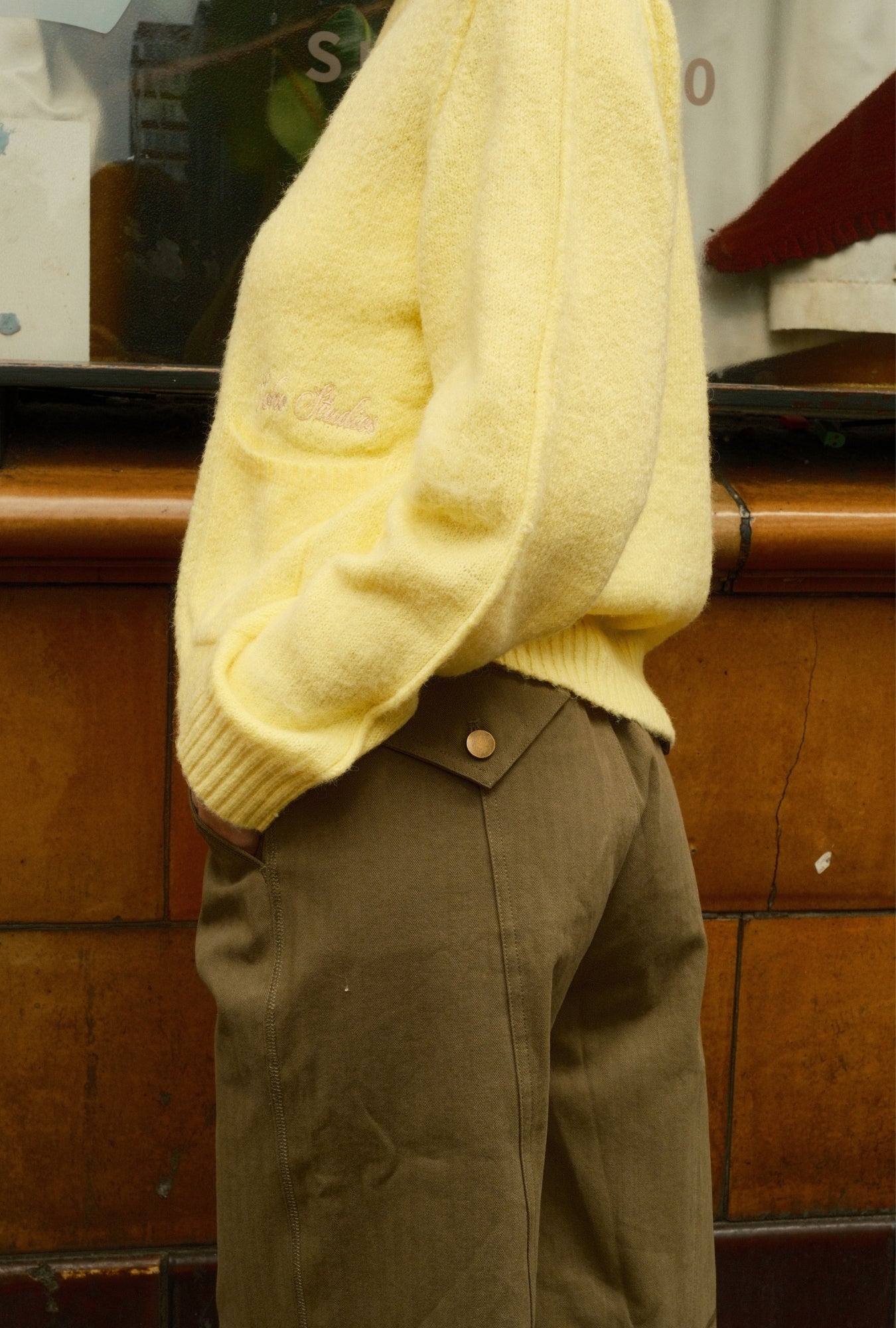 Person wearing a yellow sweater and brown pants in front of a wooden panel.