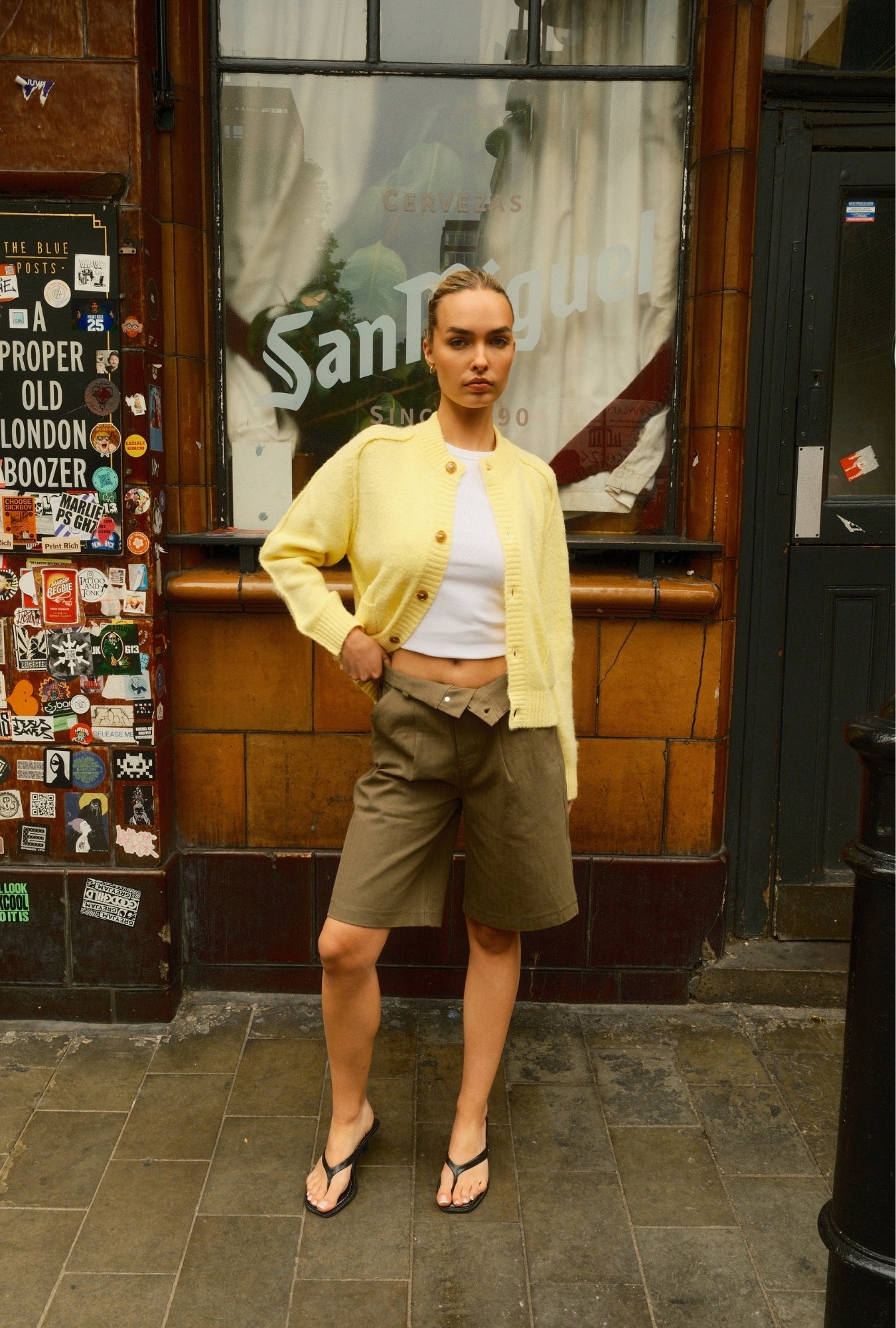 Woman in a yellow cardigan and beige shorts standing in front of a store with a glass door.