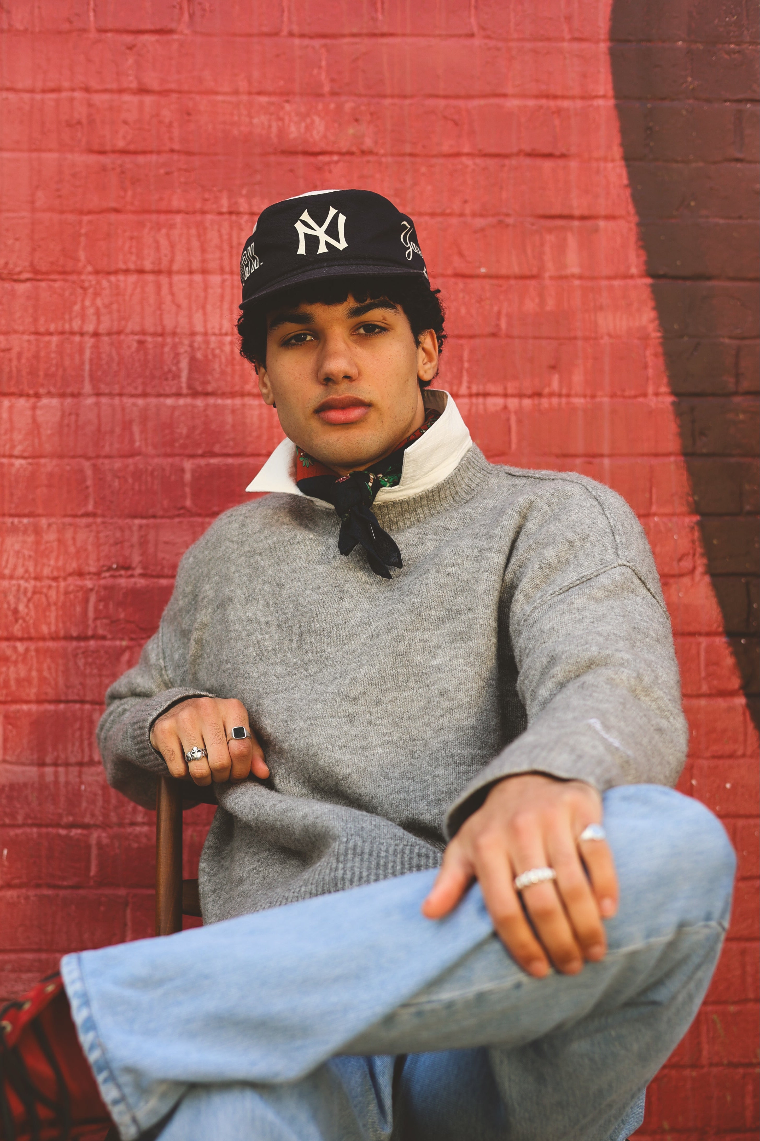 Person wearing a gray sweater and black cap against a red brick wall