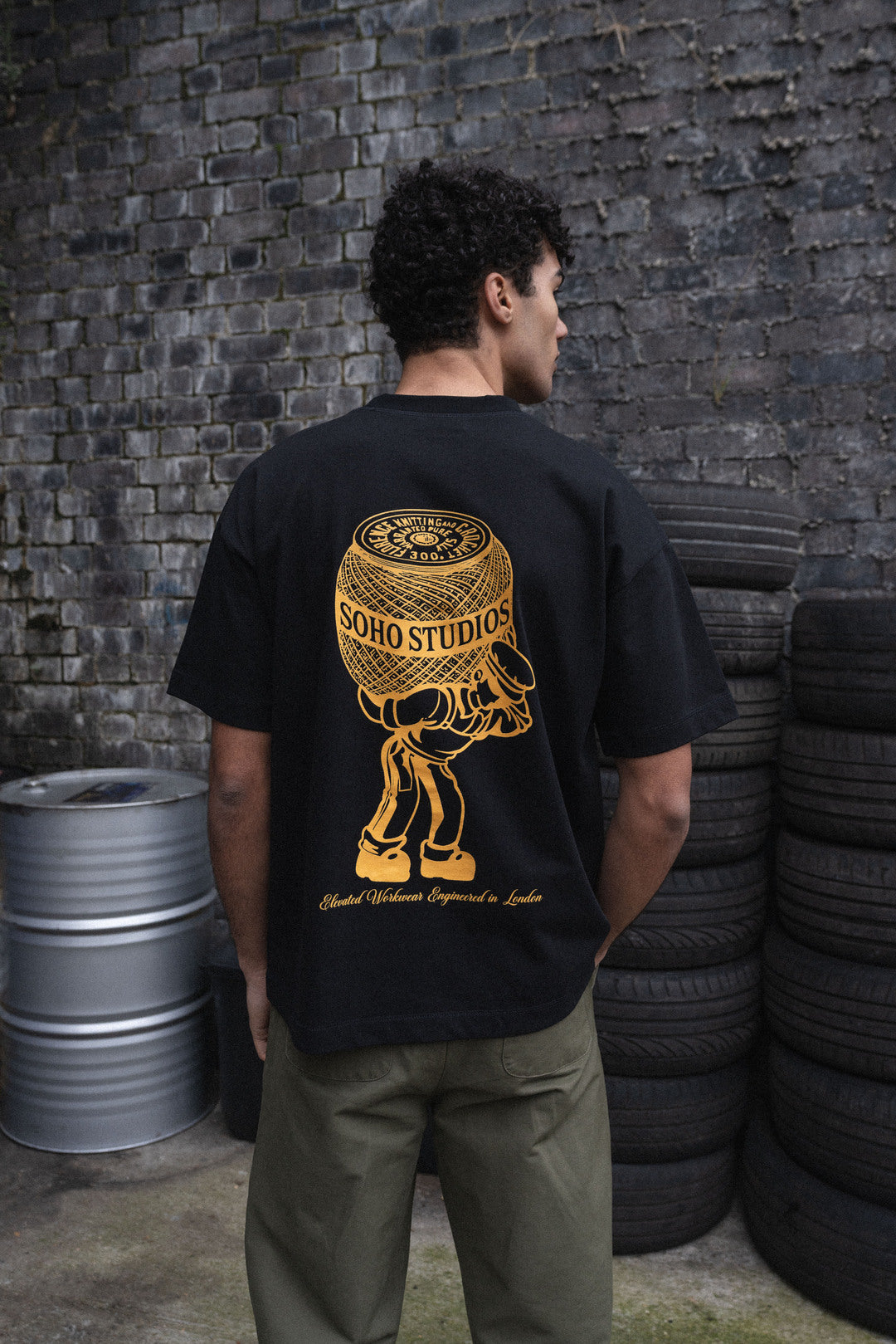 Men wearing Black 'Atlas Mascot' t-shirt with yellow screen print on the back, displaying a stylized character and the text 'SOHO STUDIOS'.