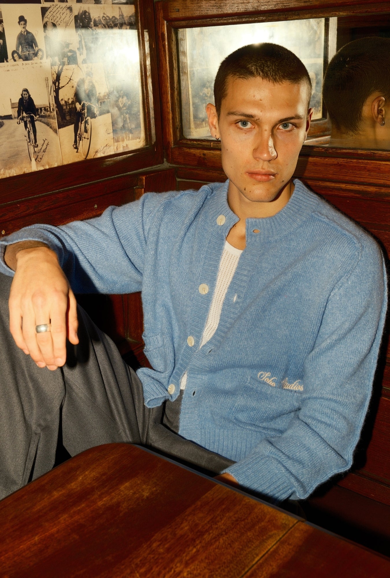 Man wearing a blue cardigan sitting in a room with wooden furniture and framed pictures on the wall.