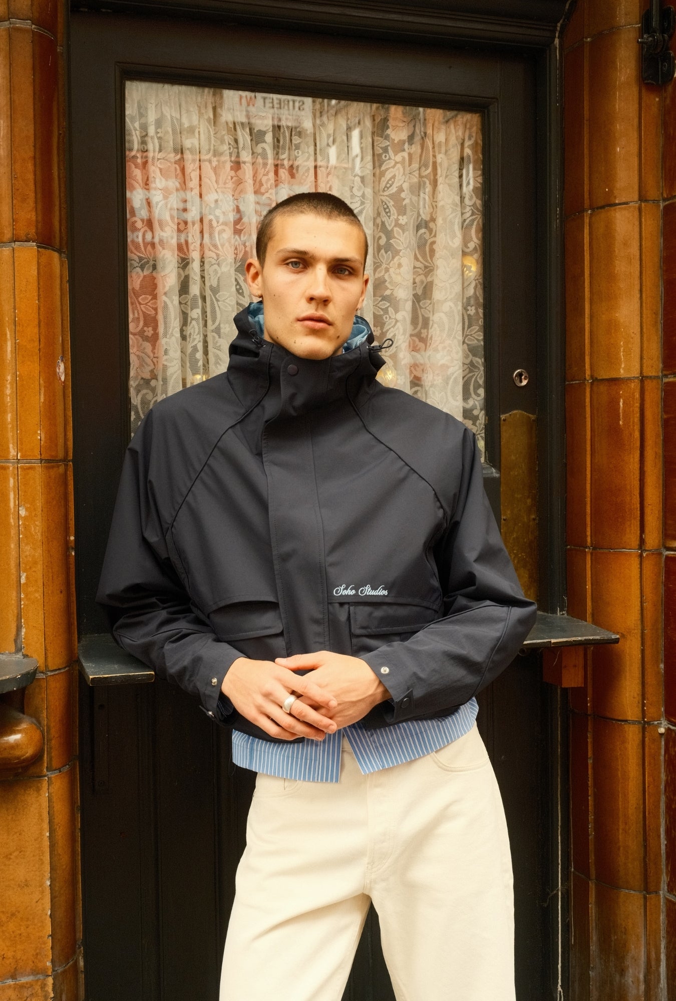 Man wearing a dark jacket with a brand logo, standing in front of a textured wall.