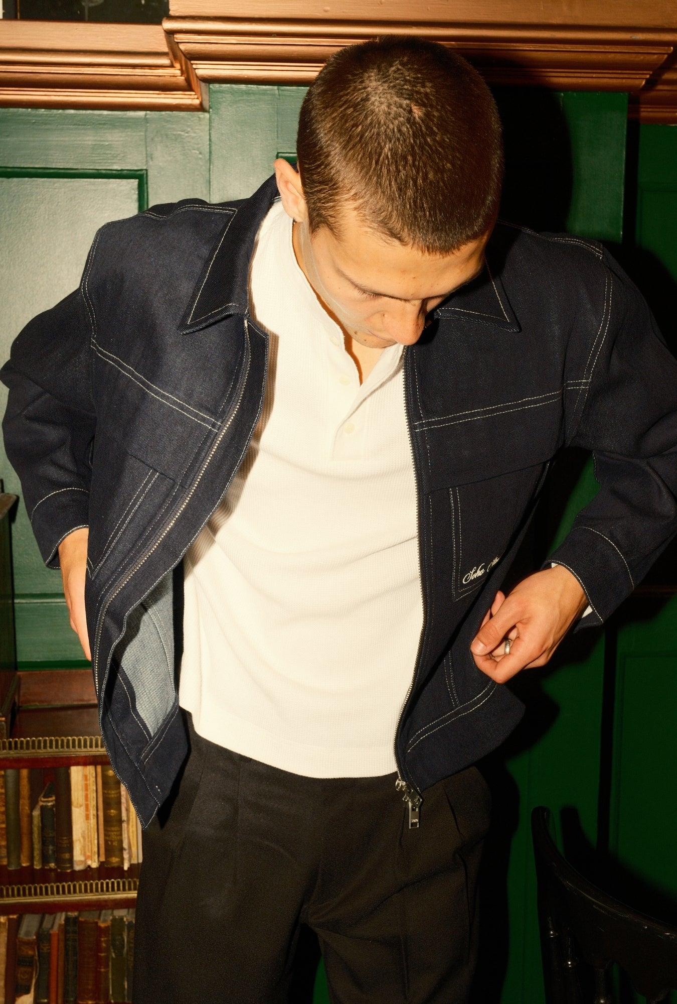 Man wearing a dark blue jacket over a white shirt in a room with green walls and bookshelves.