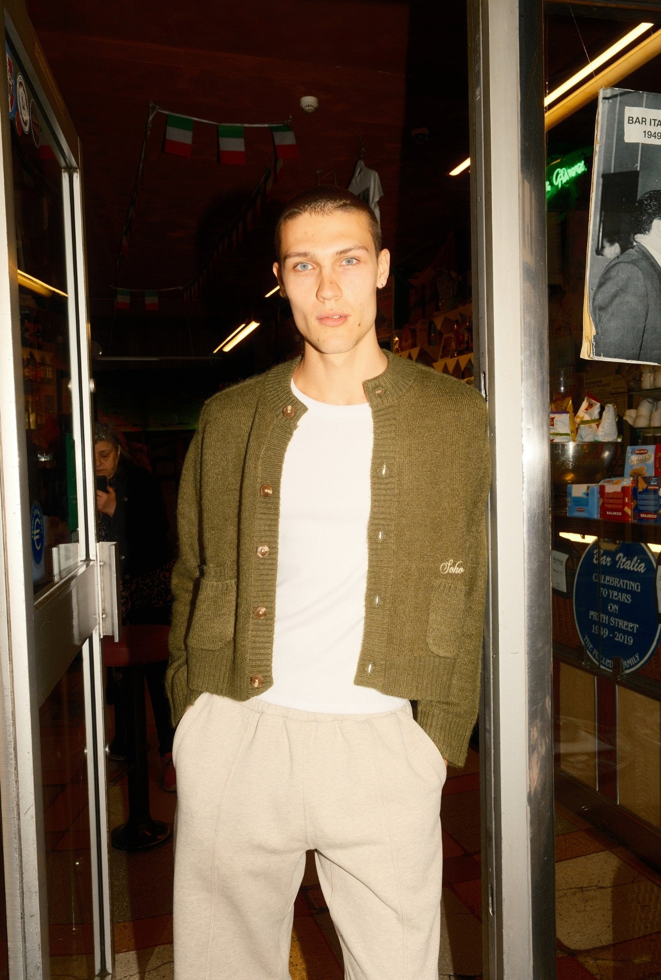 Man standing in a doorway wearing a green jacket and white shirt.
