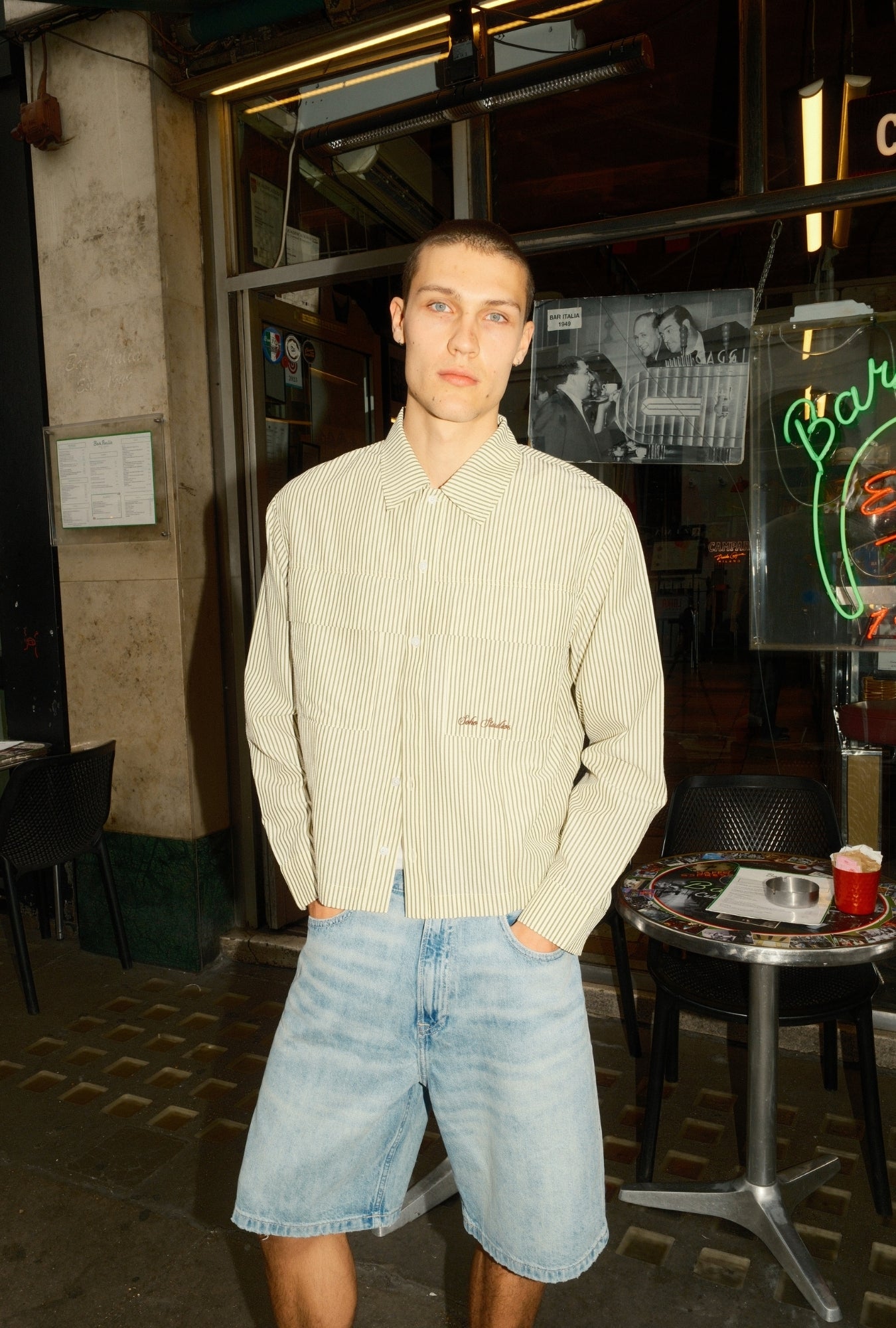 Man standing outside a bar with neon signs and patrons inside.