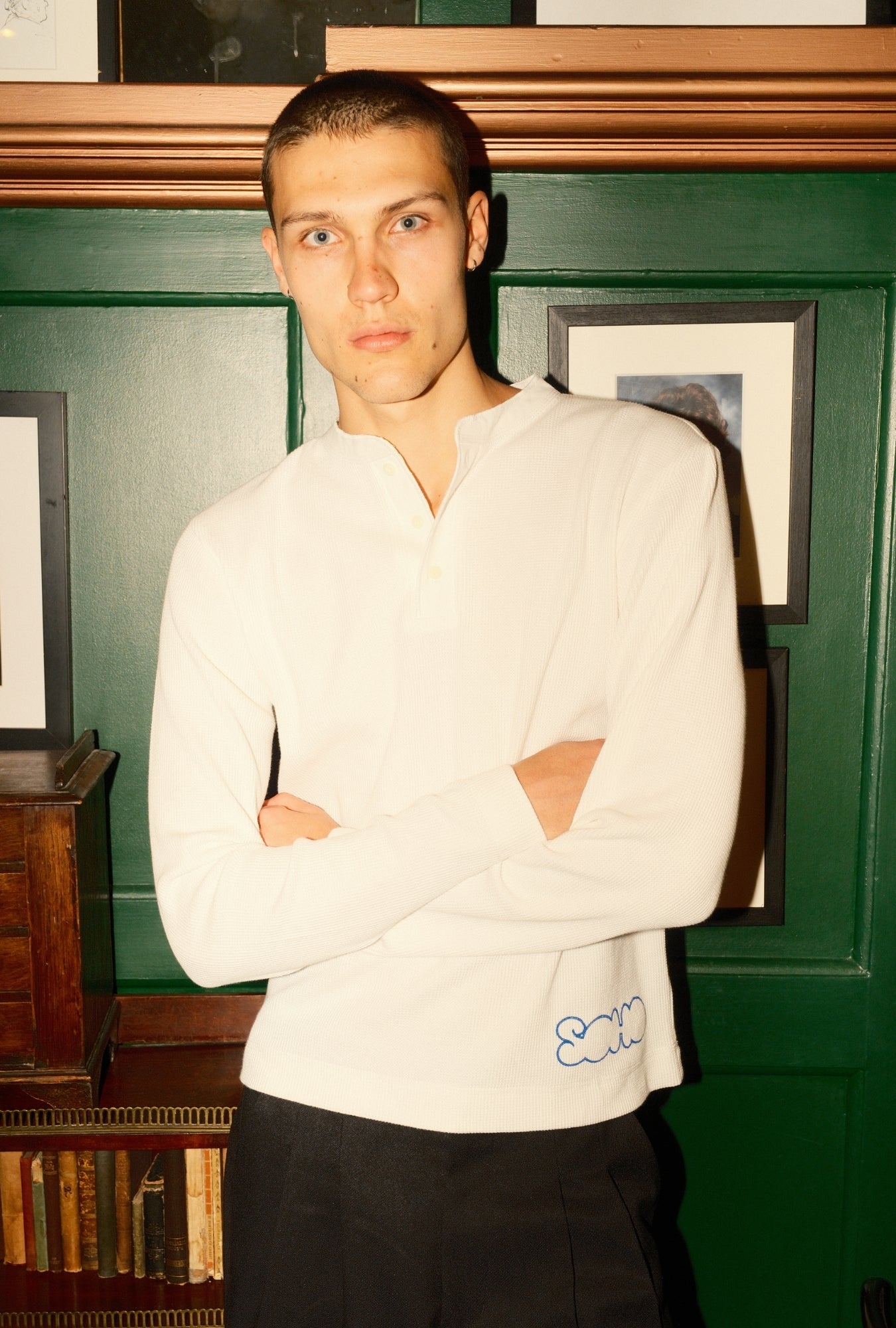 Man wearing a white sweater with a logo, standing in a room with green walls and framed pictures.