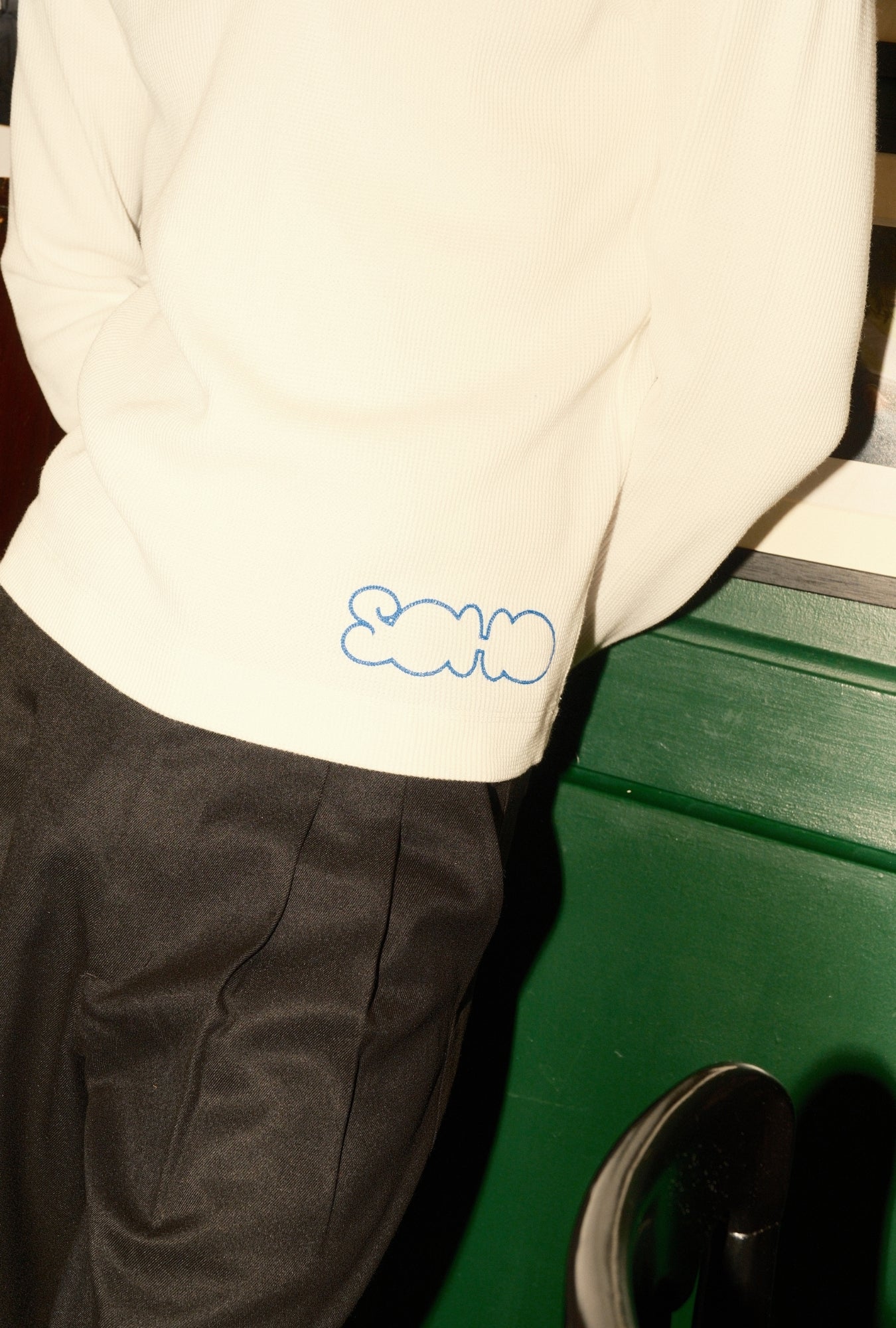 Person wearing a white sweater with a blue logo, standing next to a green vehicle.