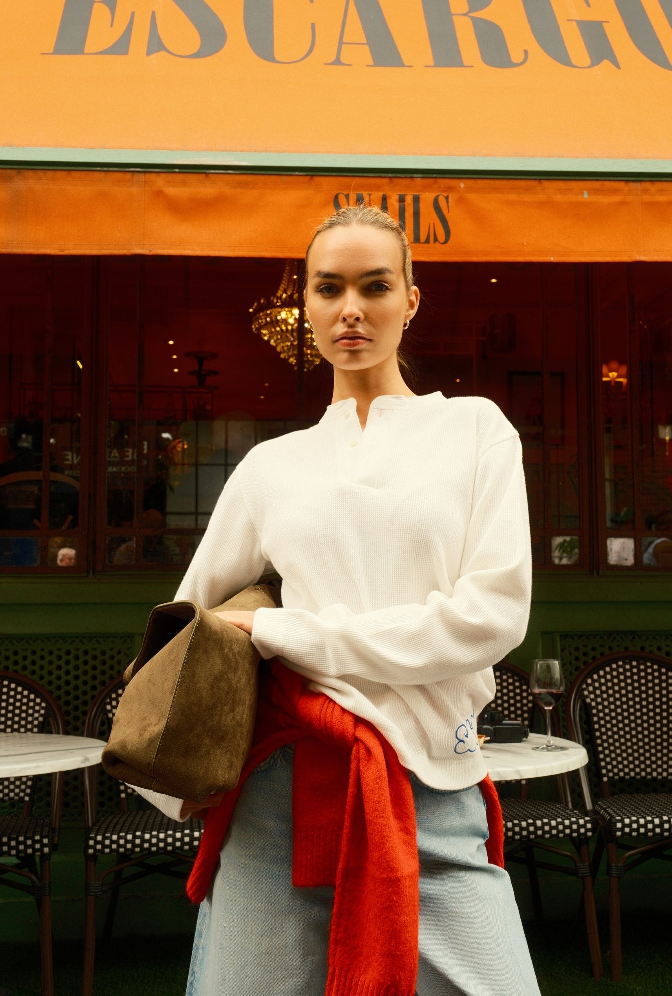 Woman in a white sweater with a red scarf standing in front of an 'Escargot' restaurant.