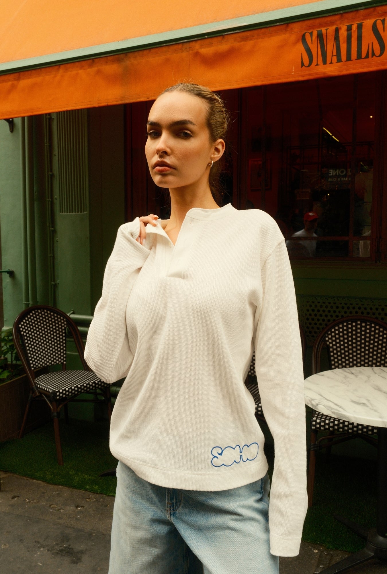Woman wearing a white sweater with blue text standing in front of a building with an orange awning.
