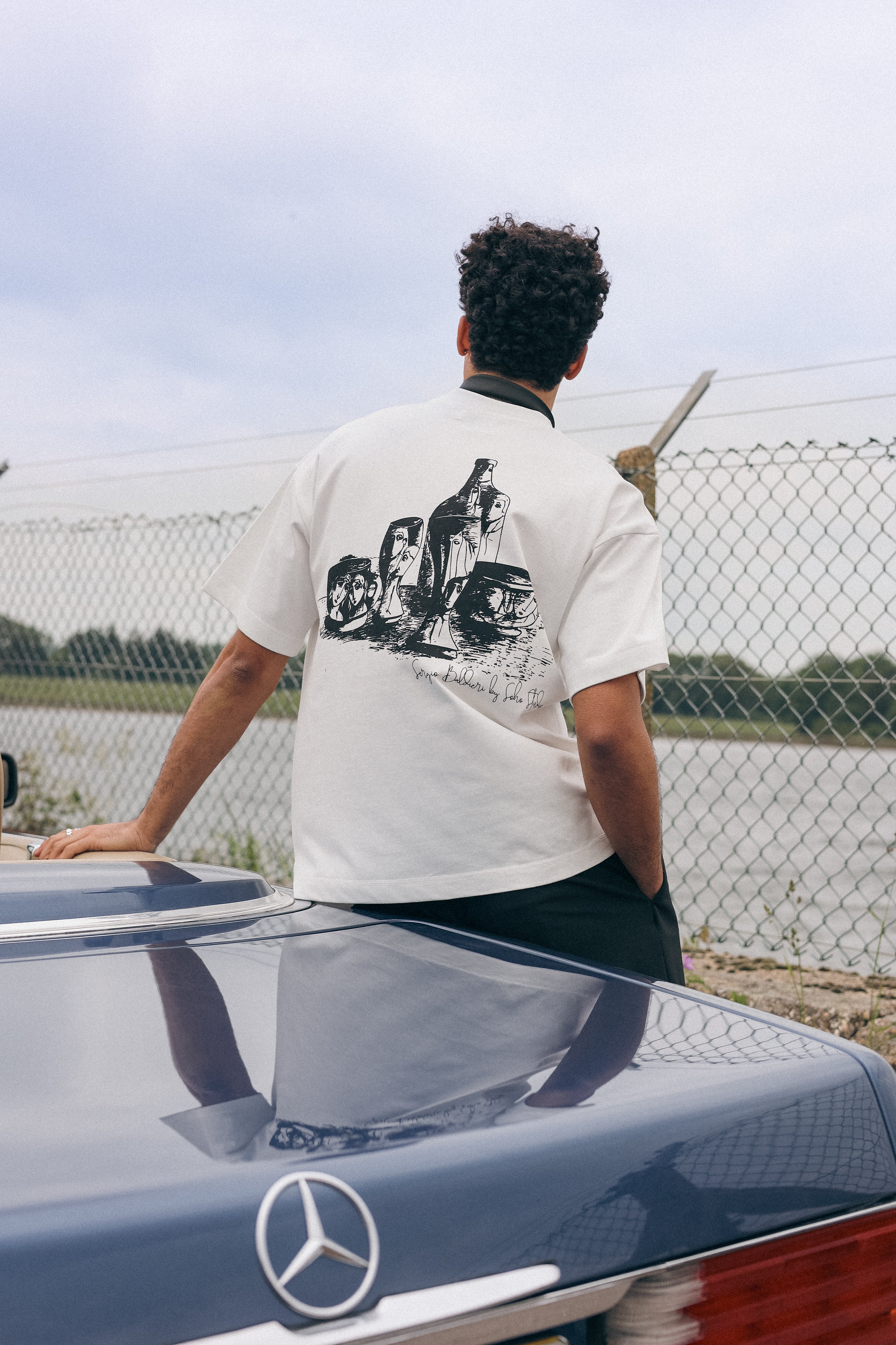 Person wearing a white t-shirt with graphic design, standing next to a Mercedes-Benz car.
