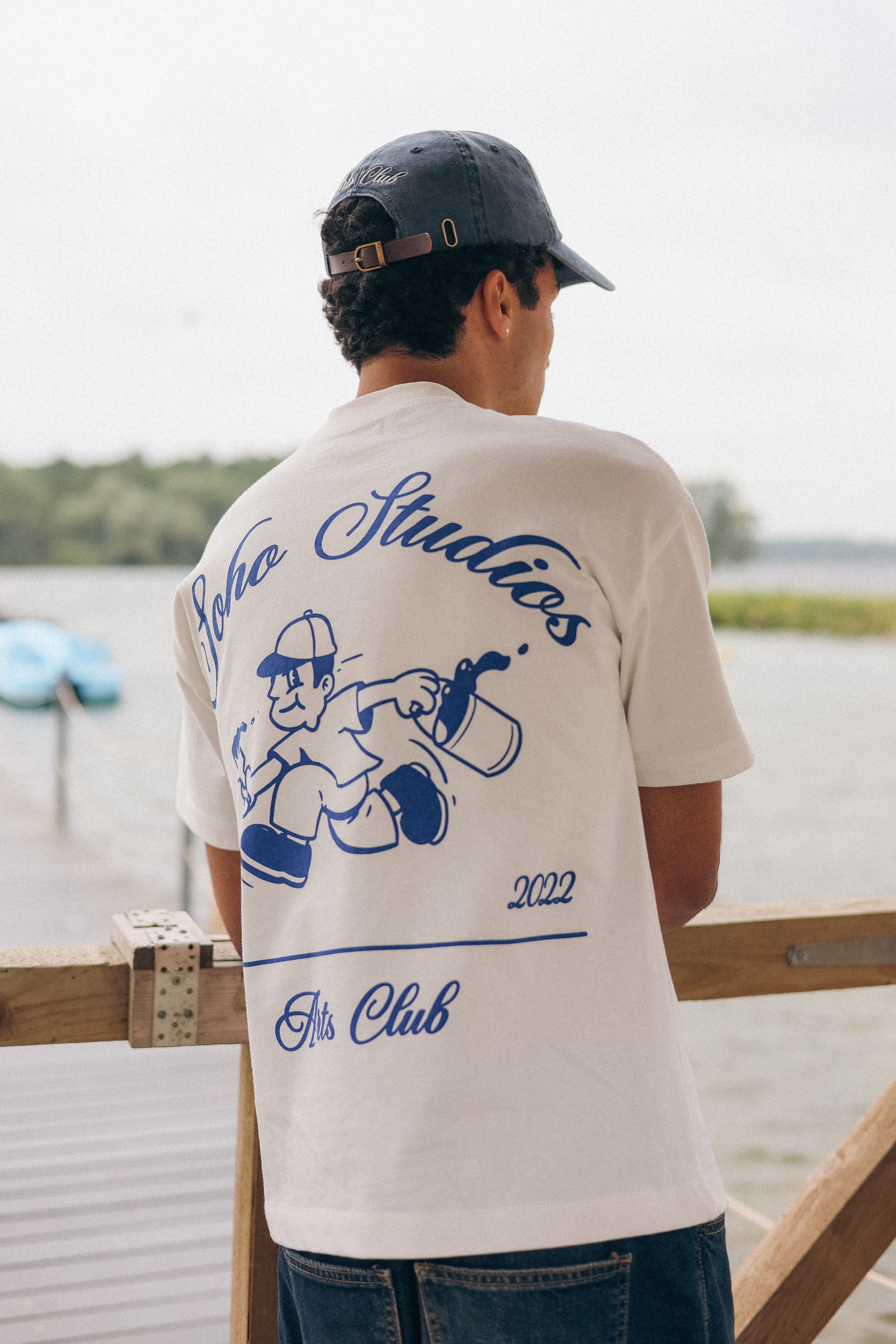 Person wearing a t-shirt with 'Soho Studios' design outdoors by a body of water.