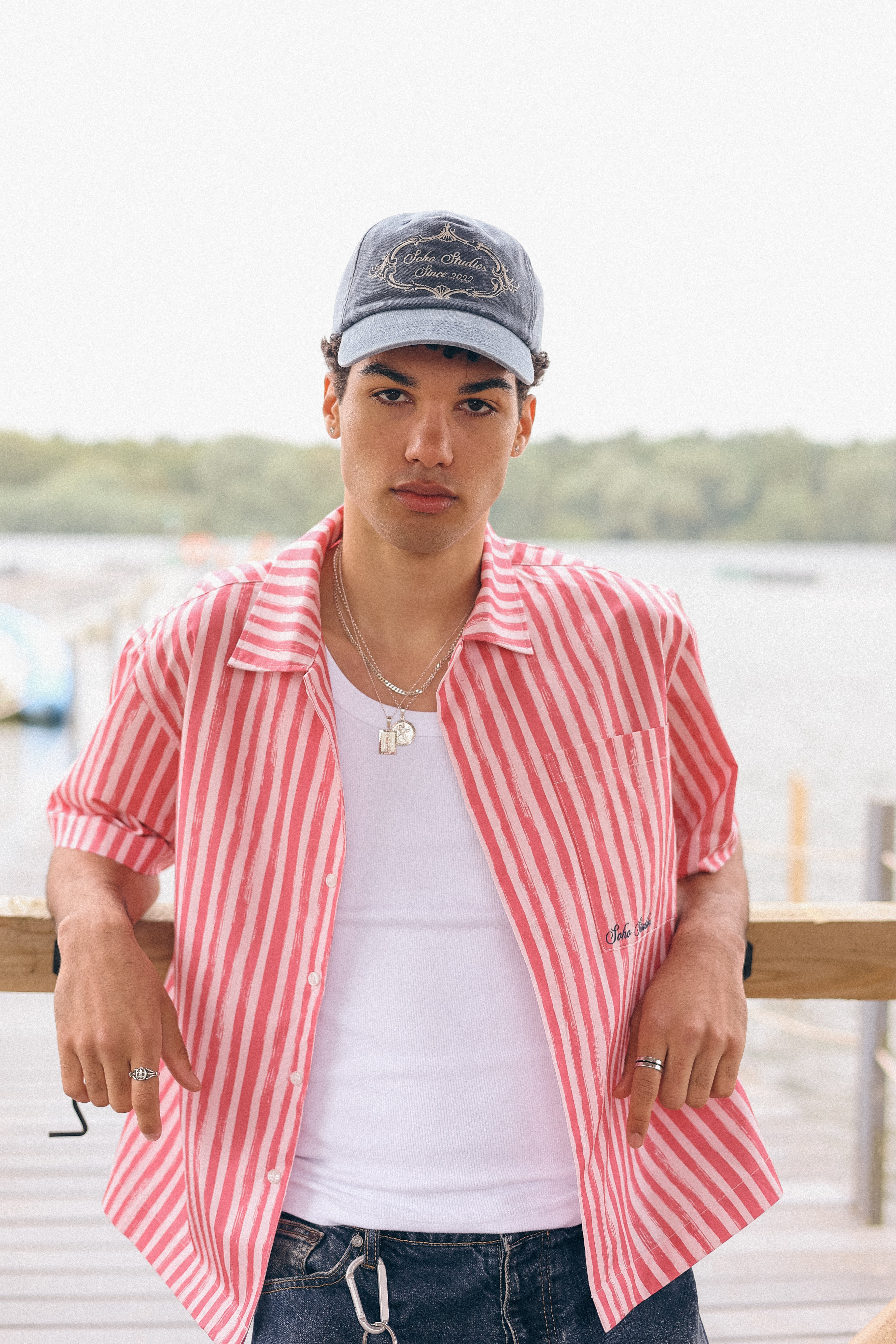 Person wearing a red and white striped shirt over a white t-shirt, standing outdoors with water and greenery in the background.