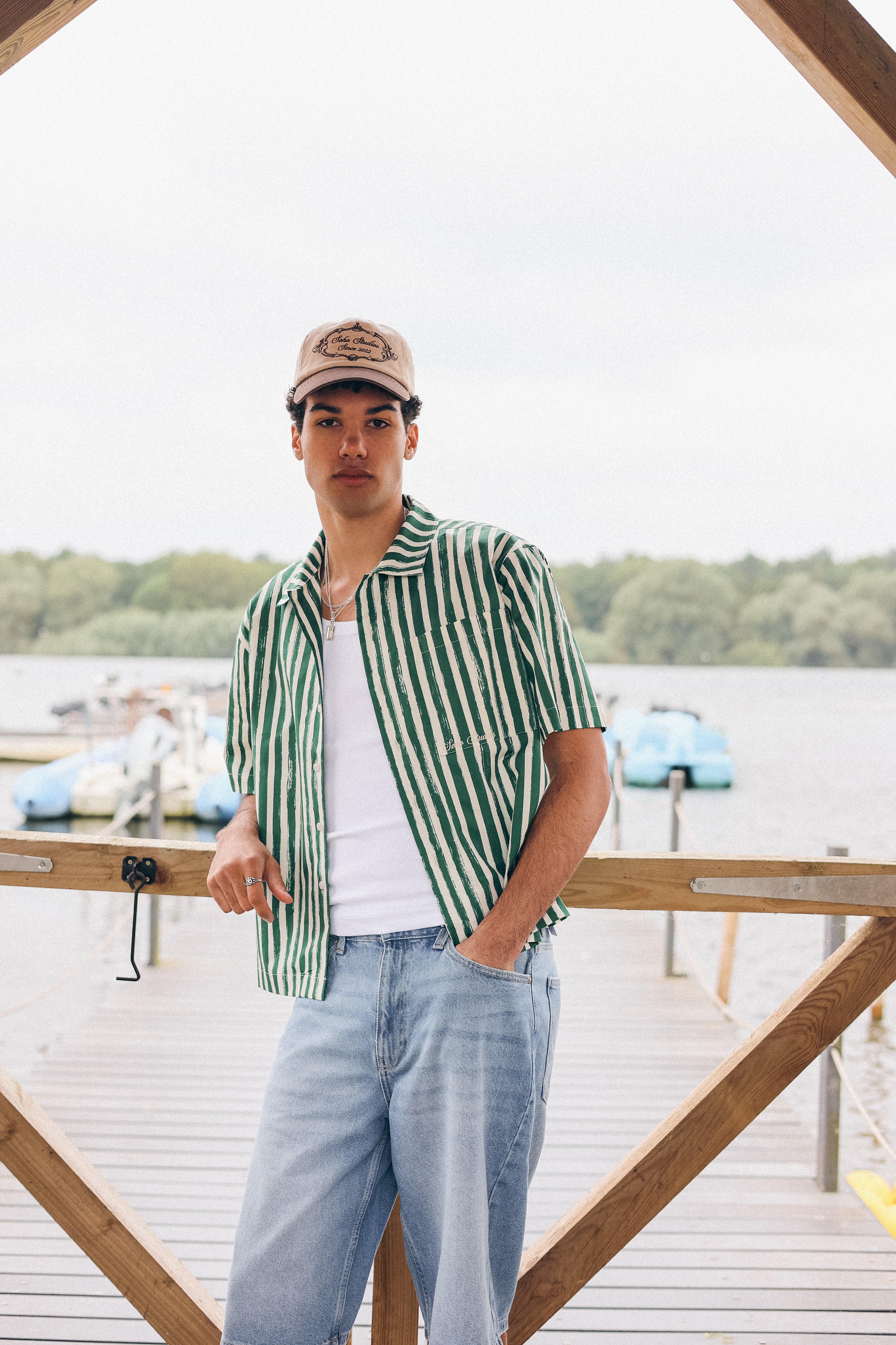 Man wearing a green and white striped shirt and cap standing on a dock with water and boats in the background.