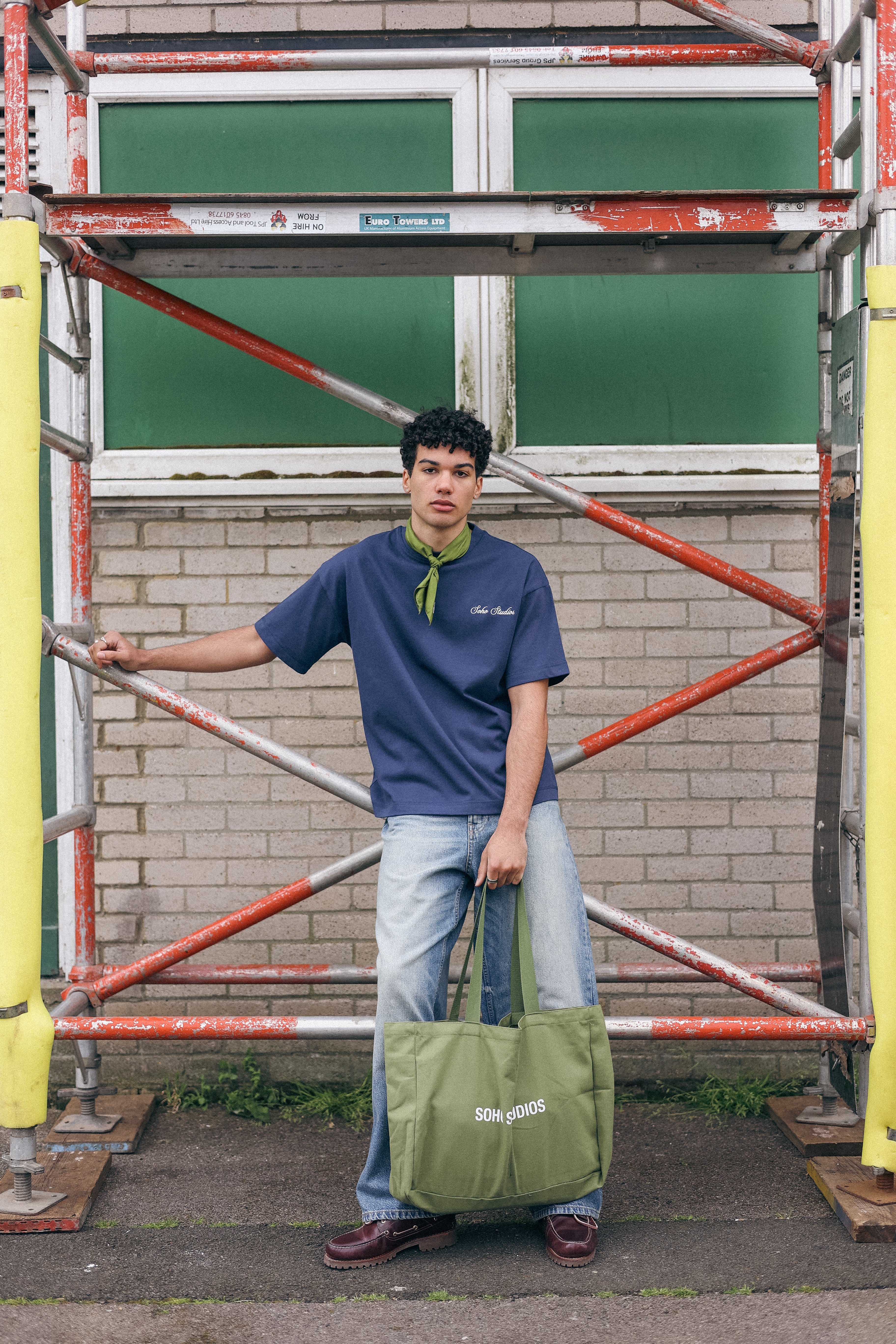 Person wearing a  navy-colored crew neck t-shirt holding a green bag in front of scaffolding
