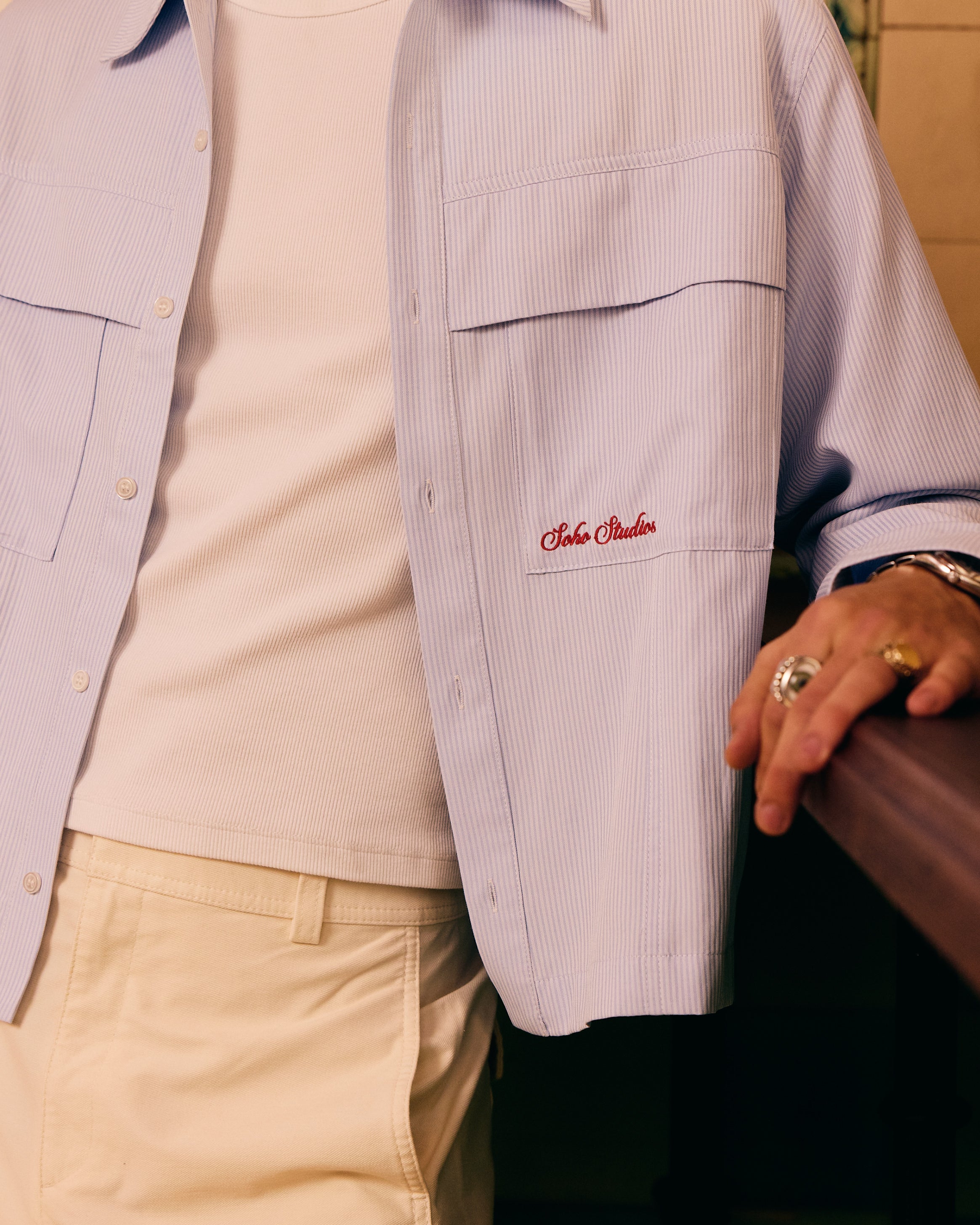 Person wearing a light purple jacket with a Soho Studio logo, beige pants, and holding a brown bag.