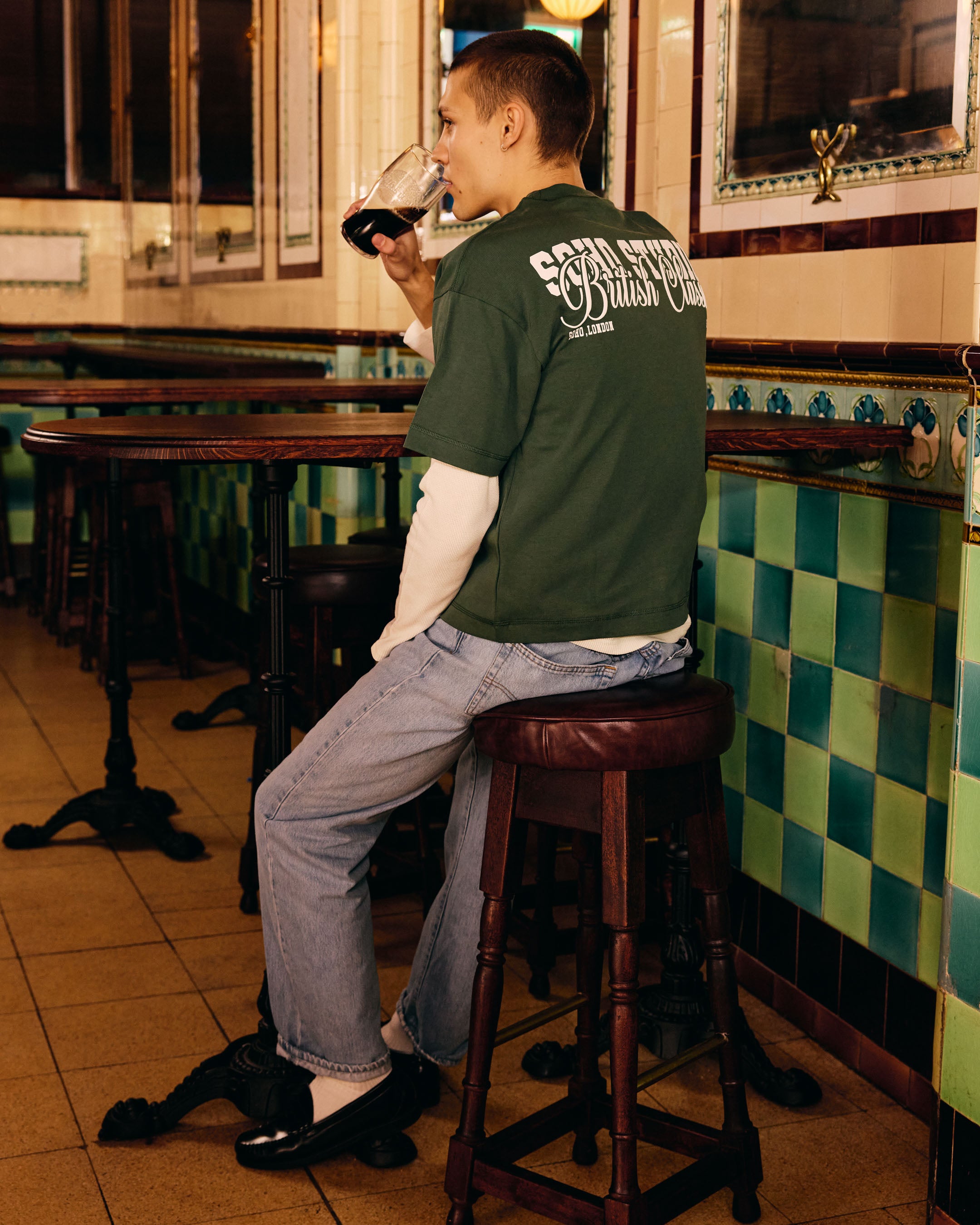 Man sitting on a stool in a bar, wearing a green t-shirt with text, holding a glass of red wine.
