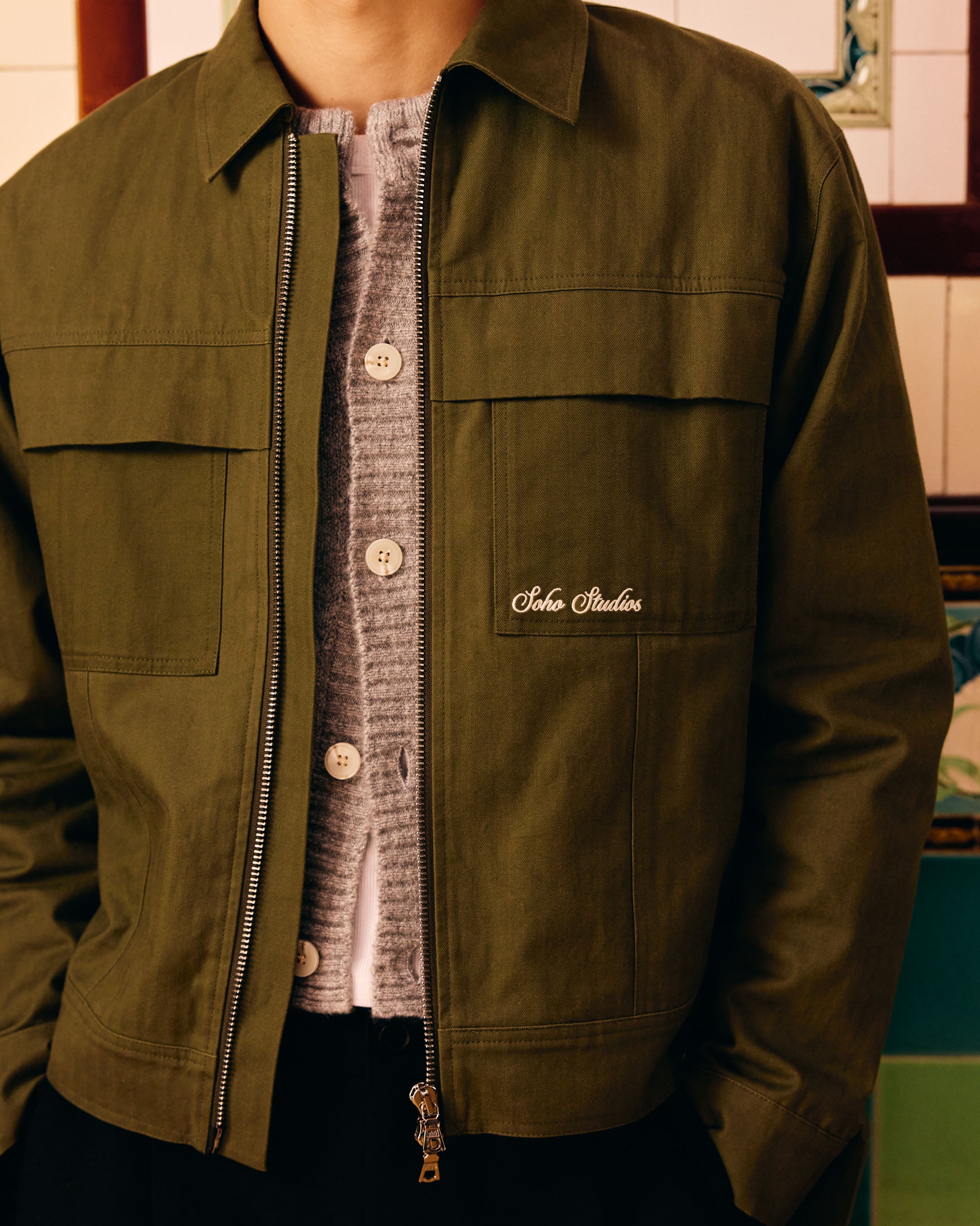 Green jacket with a brand logo worn by a person, against a blurred indoor background.