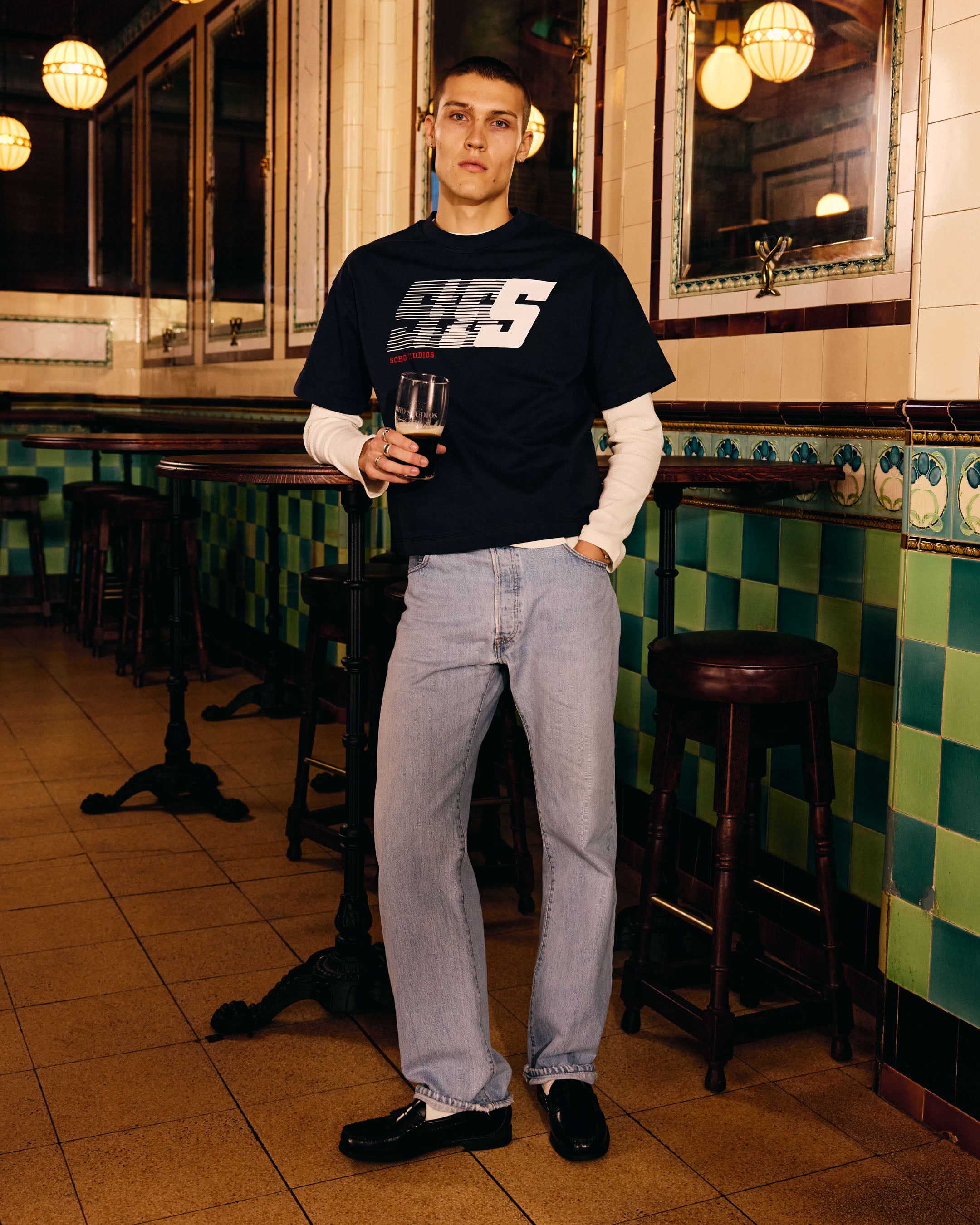 Man in a bar wearing a black t-shirt with a Soho Studio logo, holding a drink.