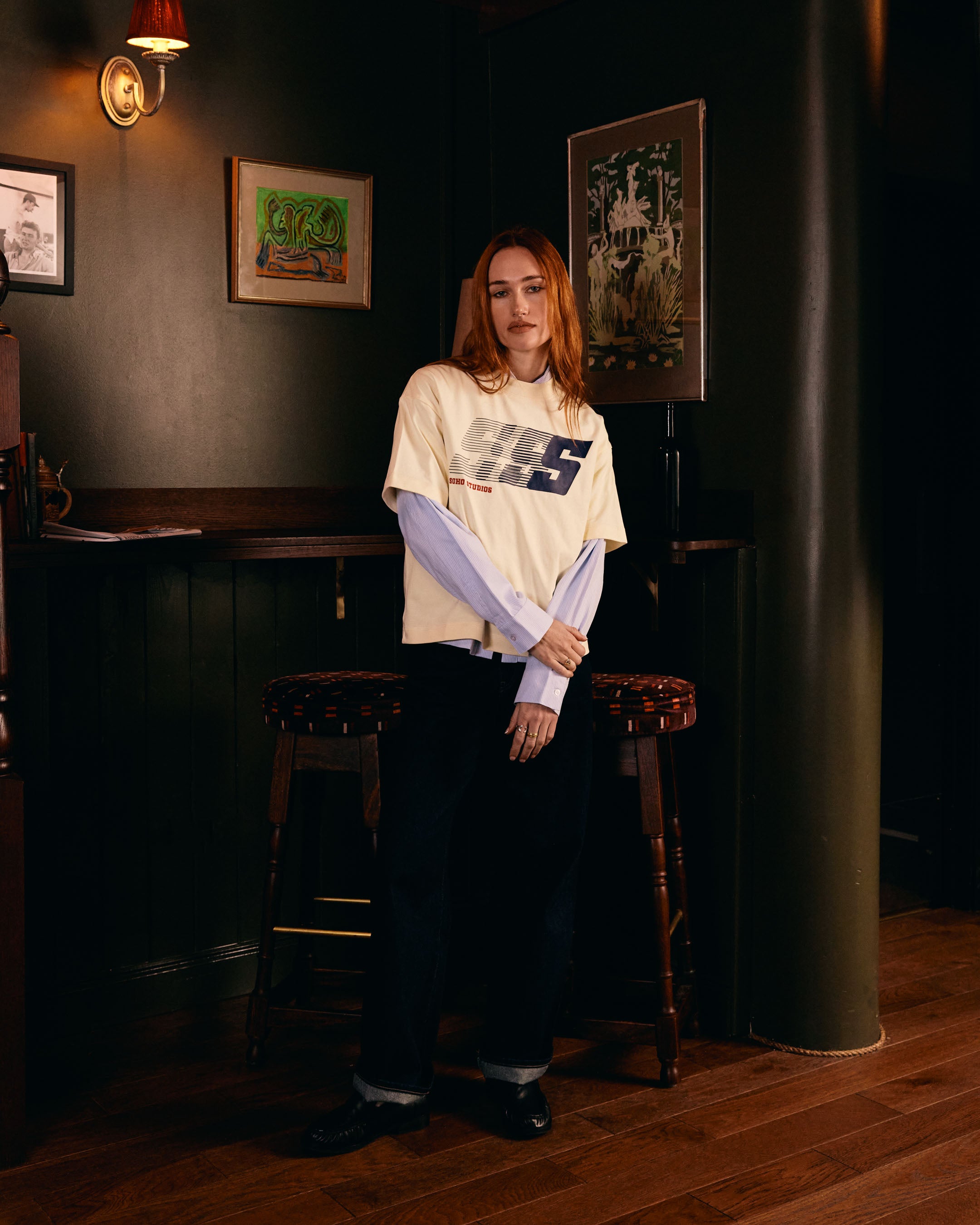 Person wearing a white t-shirt with a Soho Studio logo in a dimly lit room with wooden floors and dark walls.