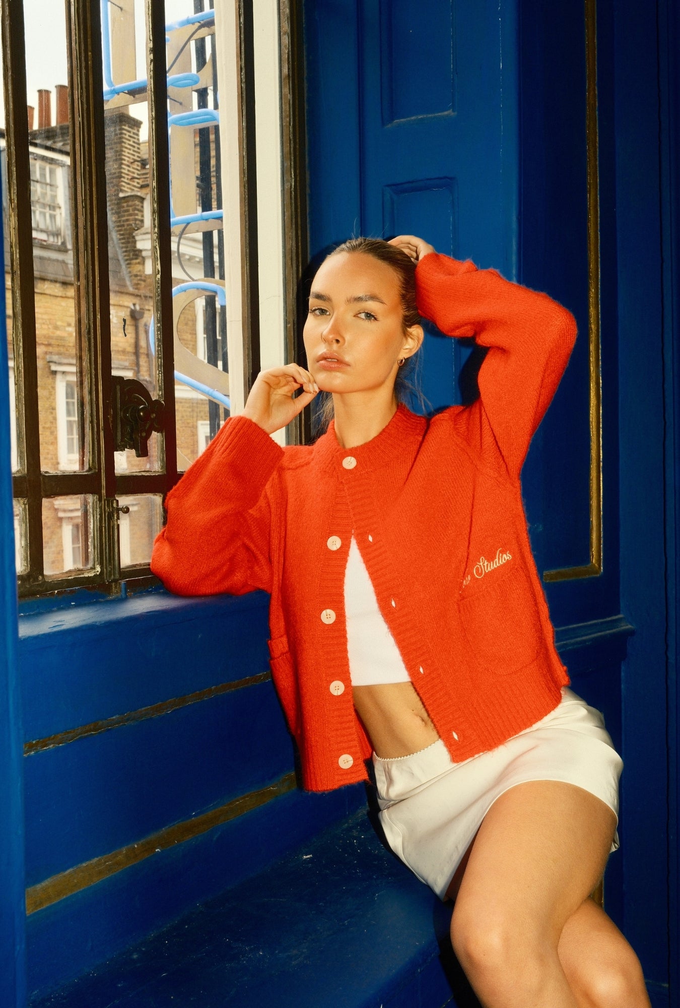 Woman in a red Soho Studio jacket sitting on a blue bench with a cityscape reflection.