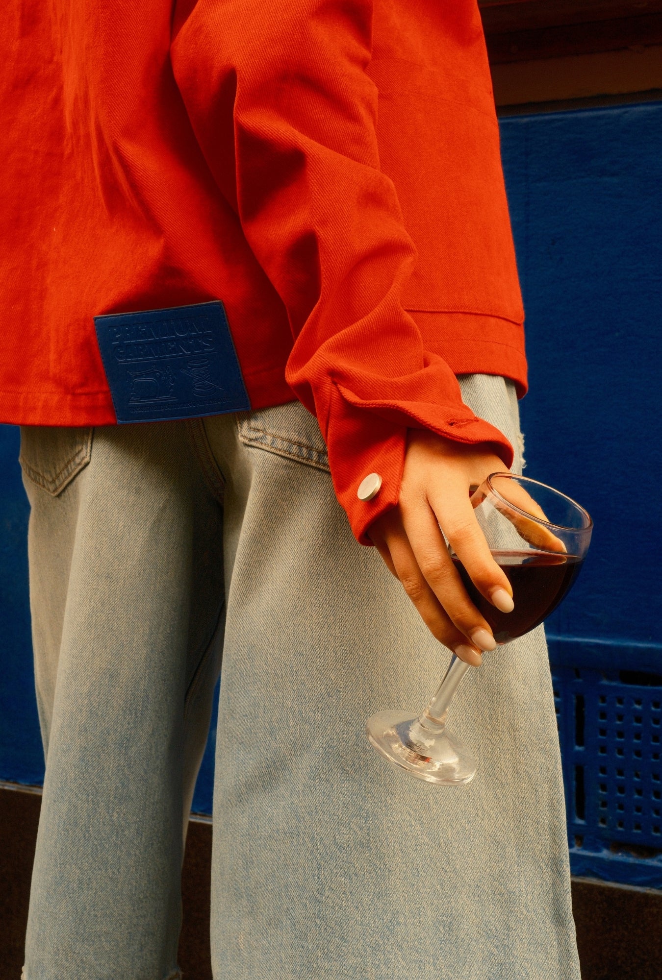 Person wearing a red jacket and light blue jeans holding a glass of red wine against a blue background