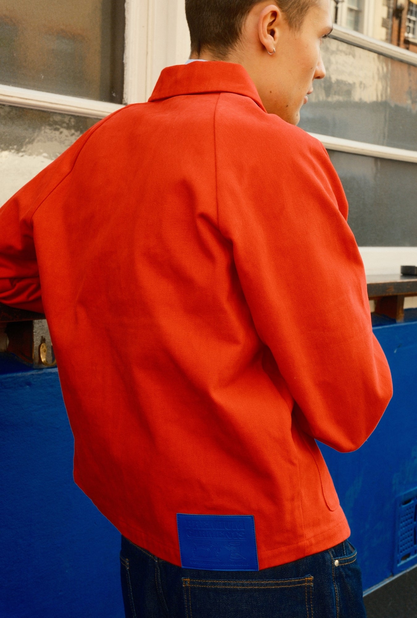 Person wearing a red jacket with a blue patch, standing in front of a blue container.