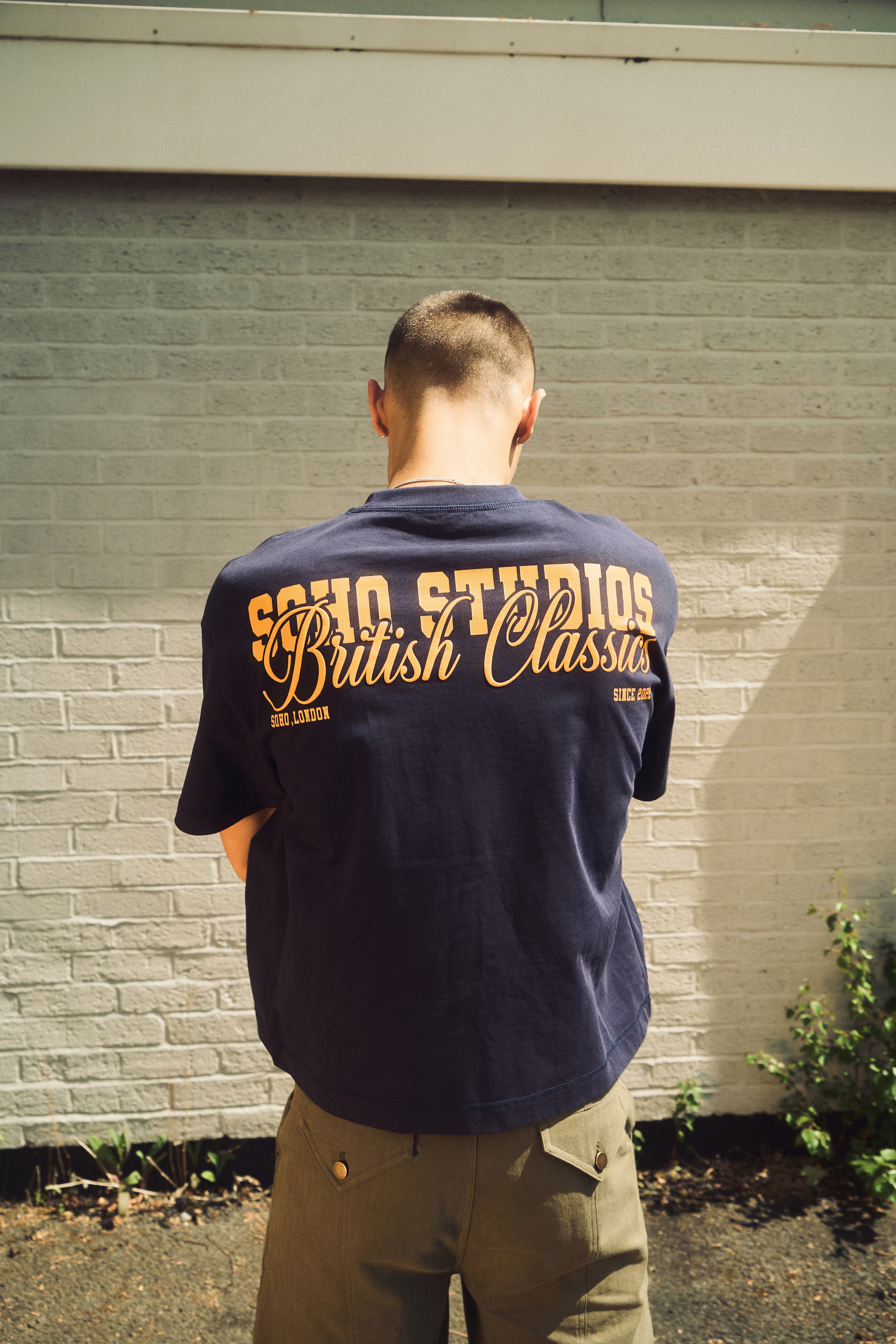 Person wearing a navy blue t-shirt with 'SOHO STUDIOS British Classics' text, standing against a brick wall.