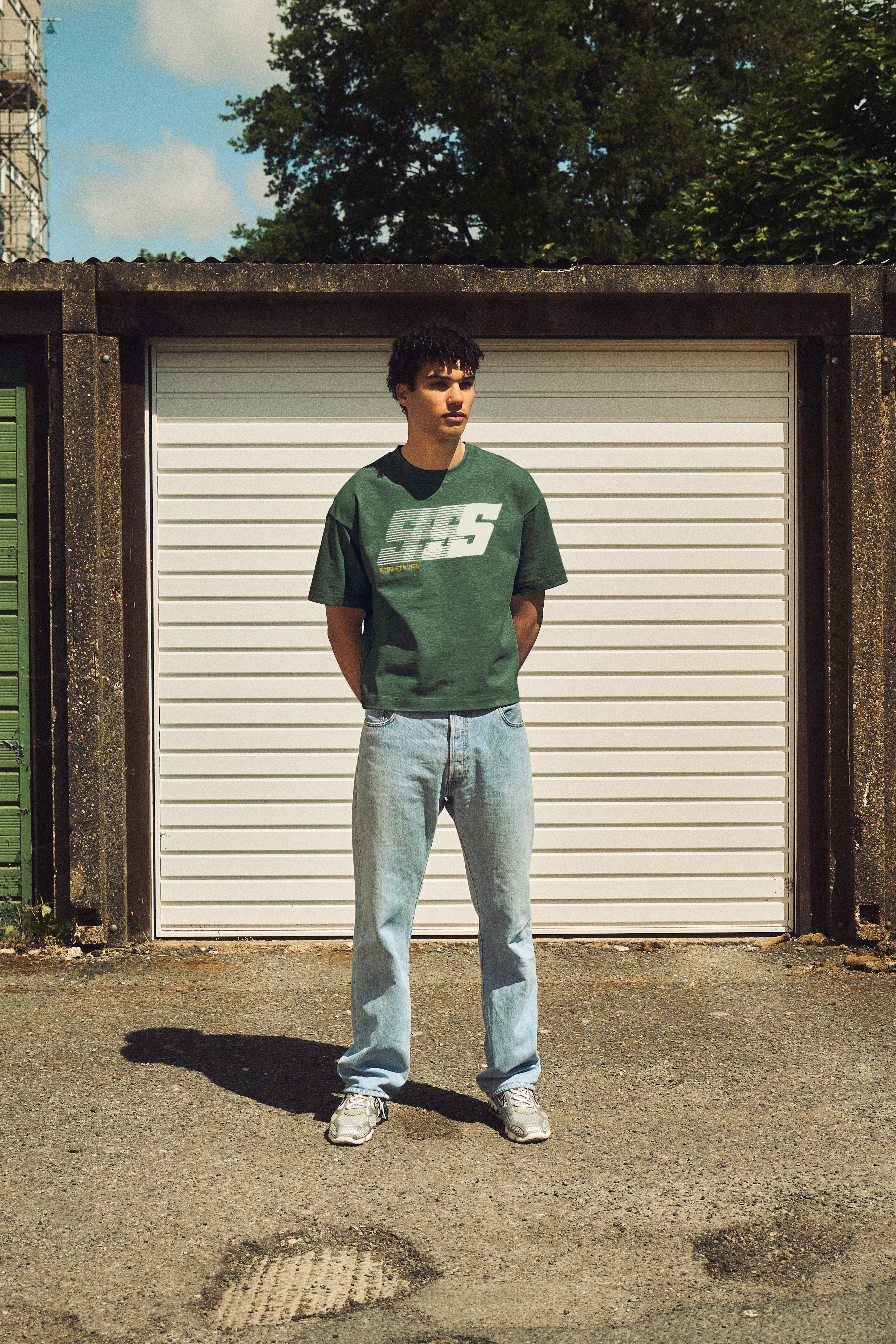 Person wearing a green t-shirt with text and light blue jeans standing in front of a white garage door.