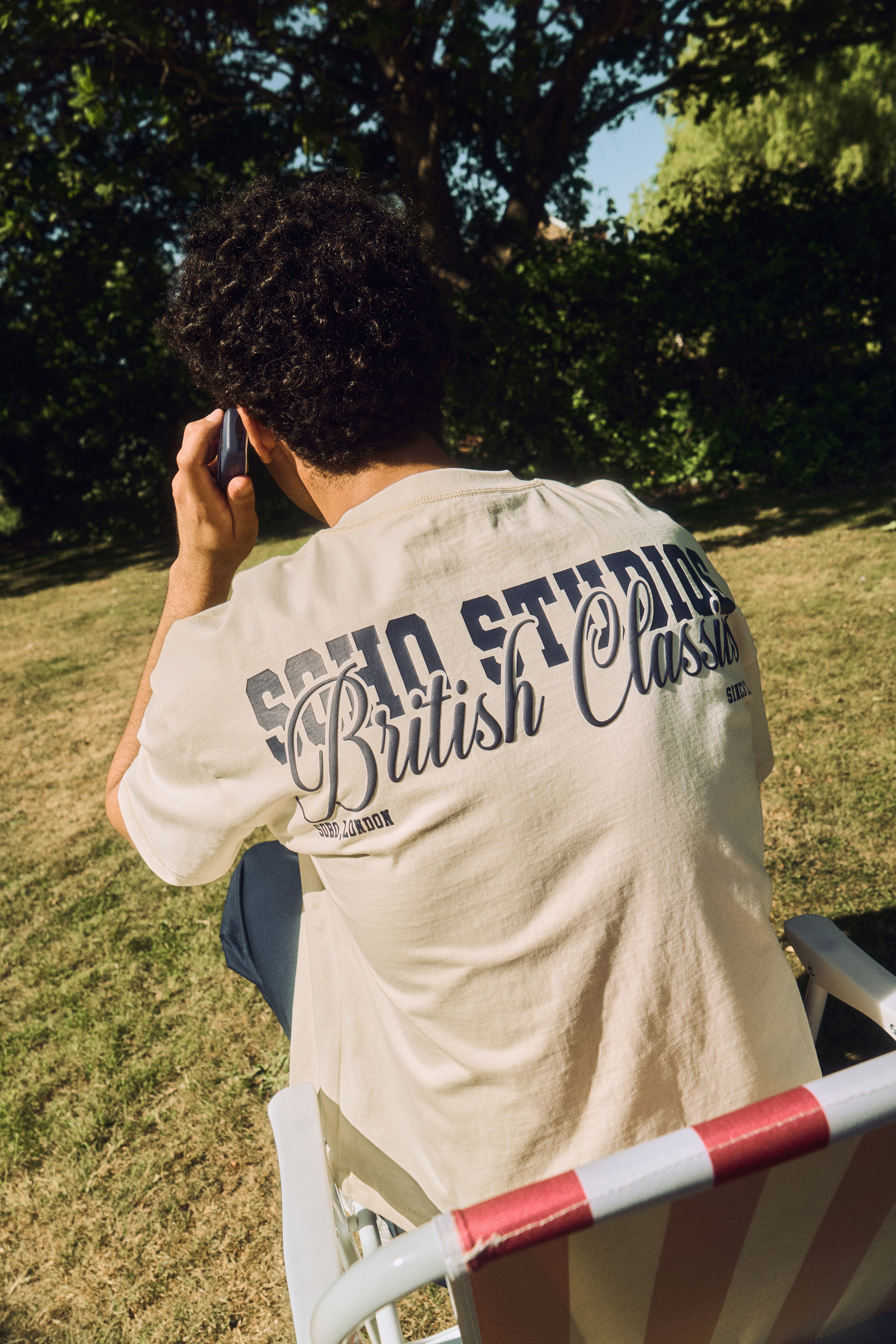 Person wearing a beige t-shirt with text, sitting outdoors on a sunny day.