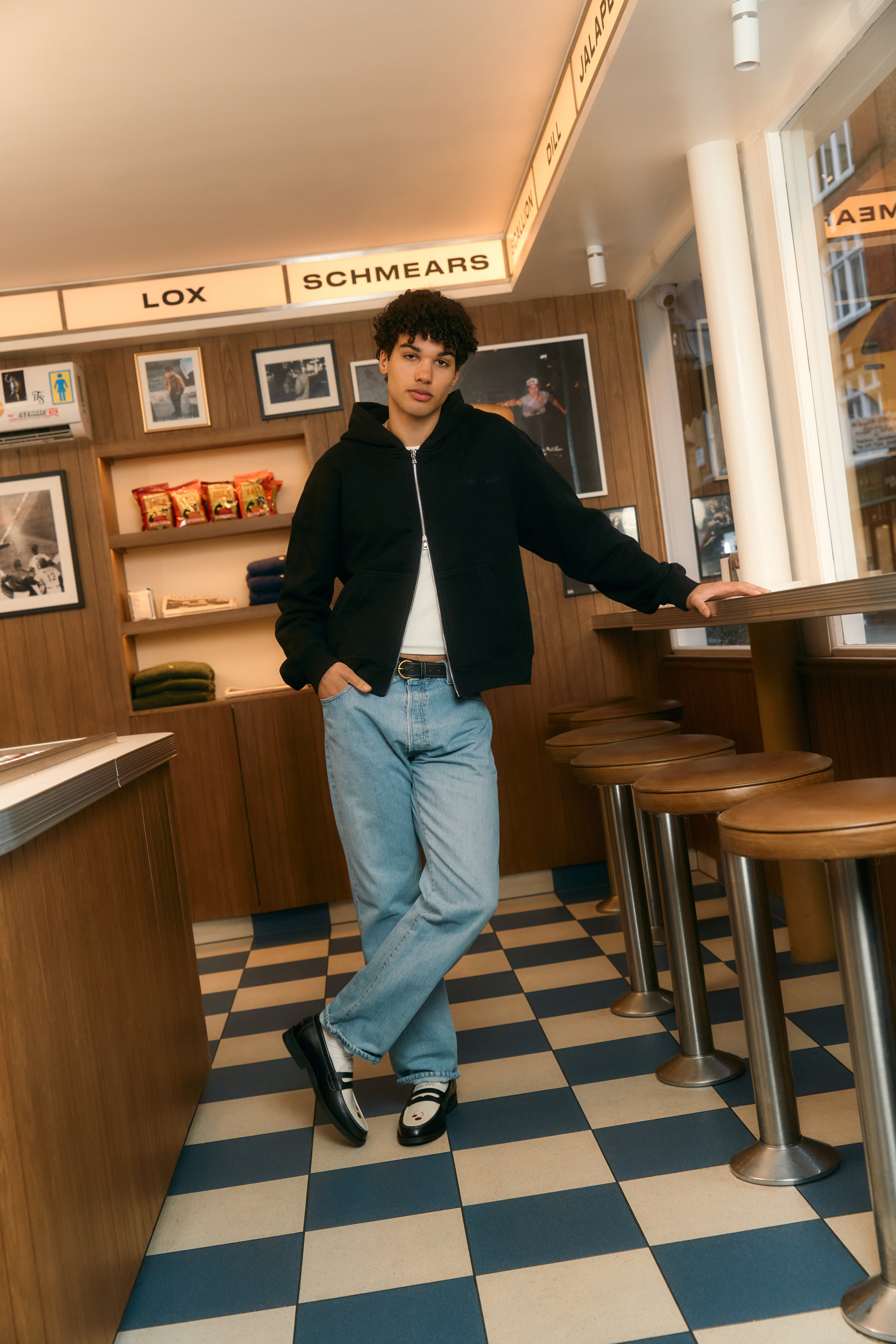 Person standing in a casual setting with checkered floor and wooden furniture